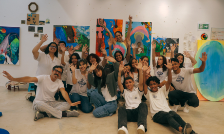 A group of smiling young people and two adults sit and kneel together in an art studio, raising their hands in celebration. Behind them, colourful abstract and figurative paintings are displayed on white walls, suggesting a collaborative art workshop or class.