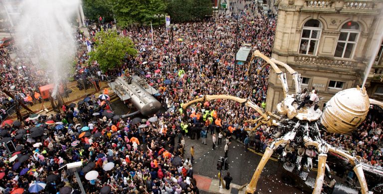 A giant mechanical spider walks through the crowded streets of Liverpool. Water shoots out of a fountain ahead.