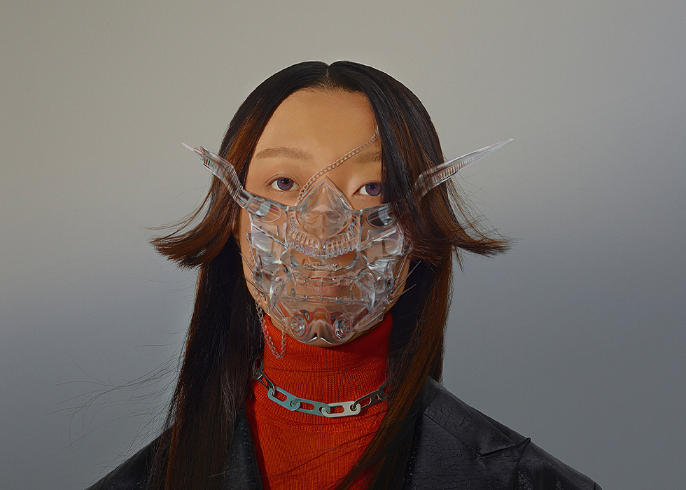 A woman with brown hair wears an abstract clear Vietnamese warrior mask with a silver chain snakinng across her face. She wears a red top with a leather jacker and chained necklace over the turtle neck of the shirt.