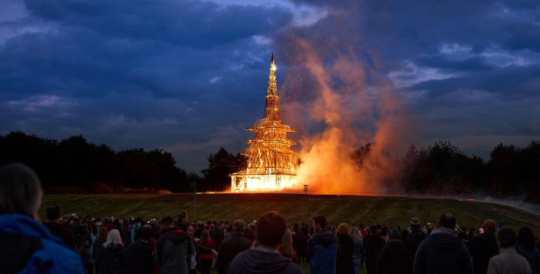 A wooden temple is engulfed in flames in the distance. Crowds watch from afar.