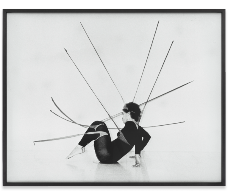 A black and white photograph with a black frame showcases a woman figure with elongated poles strapped to their body as they pose on the floor.
