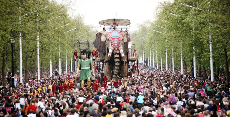 A giant elephant and girl puppet walking down the London Mall. Huge crowds of people are seen around them.