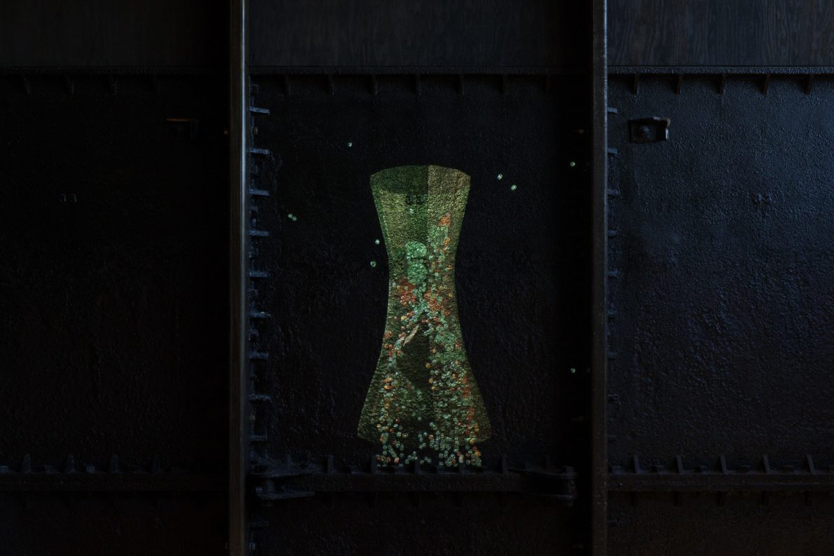 An installation of a sculpture artwork is shaped like an hourglass and holds in its green shape objects of different colours.