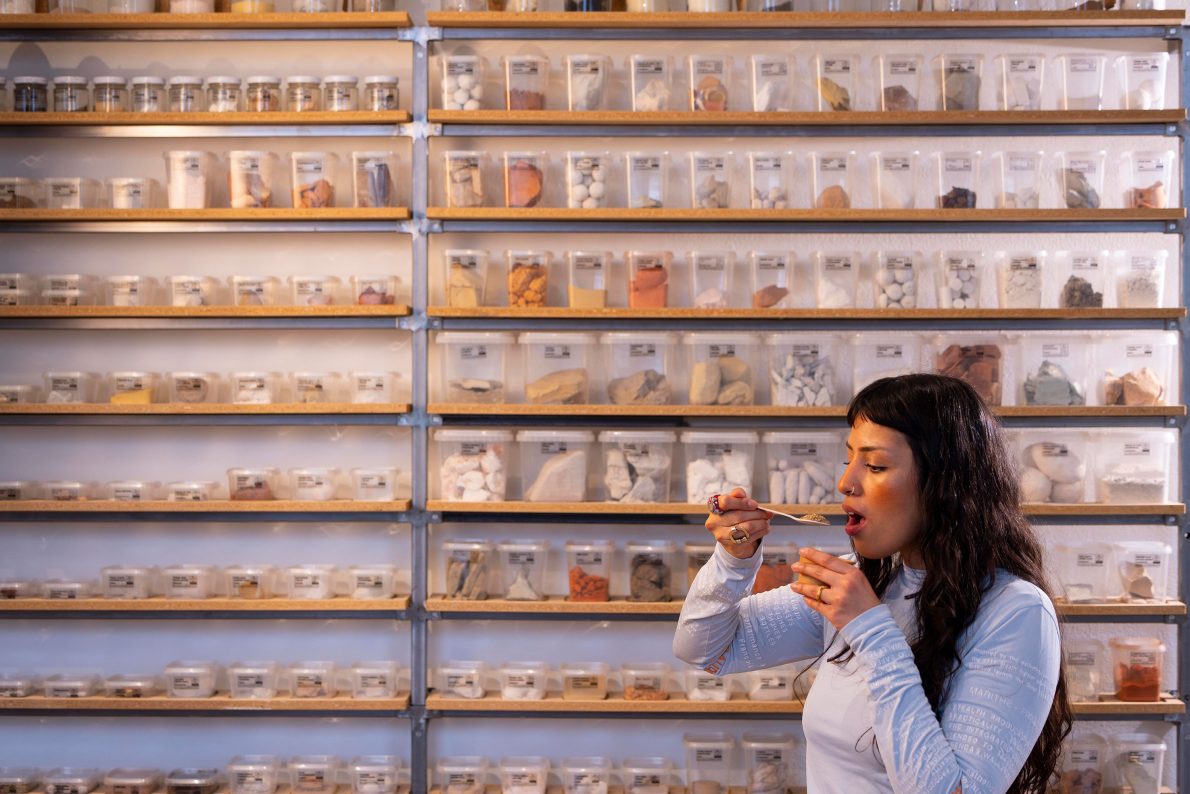 In front of a display case holding samples of earth from across the world, a woman holds a spoon to her mouth.