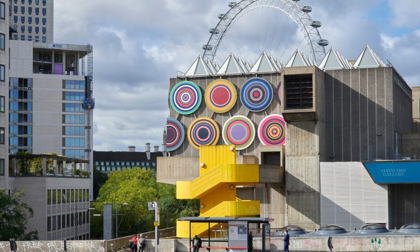 A multicoloured circular installation is on the side of a brutalist, concrete building with a striking yellow spiral staircase in front. People wait at a bus shelter as you see the London Eye and Big Ben clock tower in the background