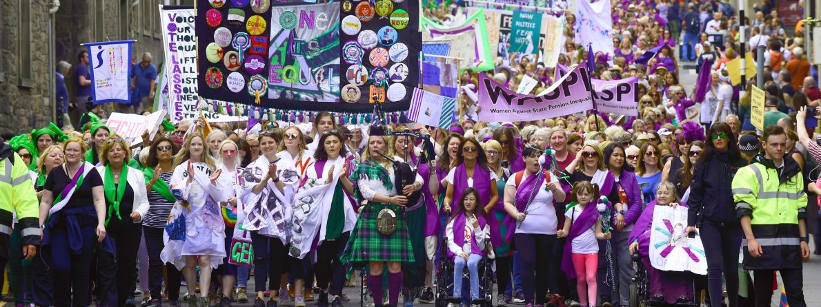 A vibrant procession with a large, diverse crowd. Women in purple, white and green sashes carry banners and signs on a city street. The scene expresses unity and empowerment.