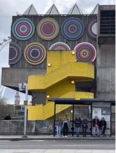 A multicoloured circular installation is on the side of a brutalist, concrete building with a striking yellow spiral staircase in front. People wait at a bus shelter as you see the London Eye and Big Ben clocl tower in the background