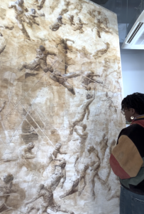 A large-scale sepia toned painting of figures moving across a featureless landscape is displayed in a gallery whilst a person