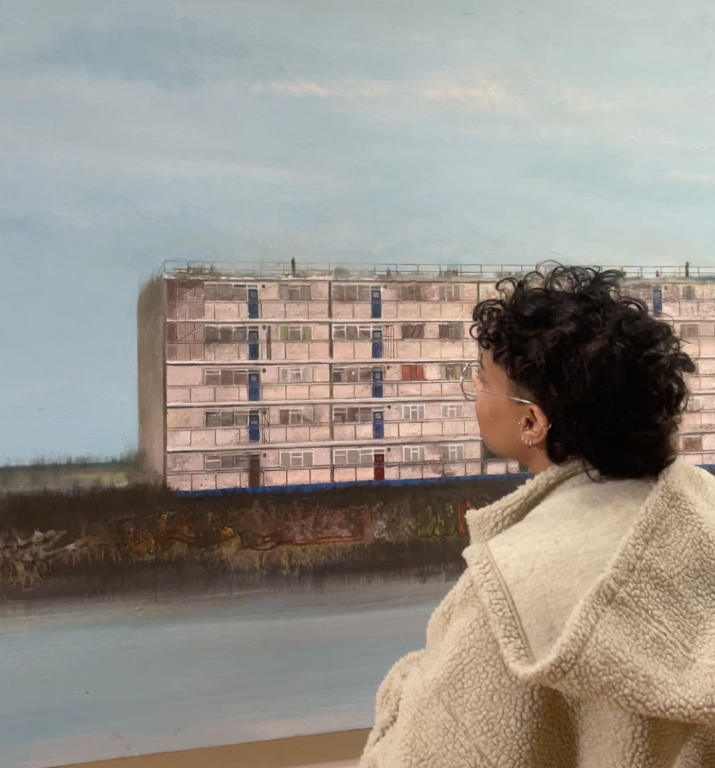 A person in a wooly jumper looks up at a large-scale painting of a block of pink flats with a pale blue background.