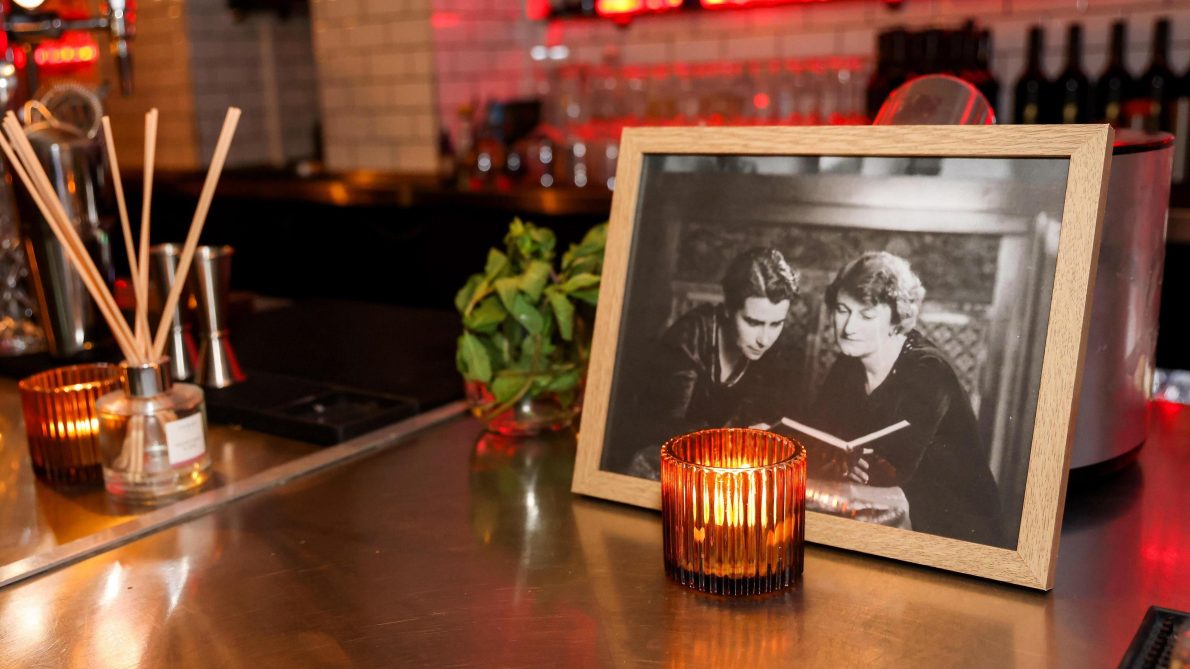 An image in a picture frame depicts two women reading a book. The pictureframe is on a table with a lit candle and incense. The atmosphere is warm and friendly.