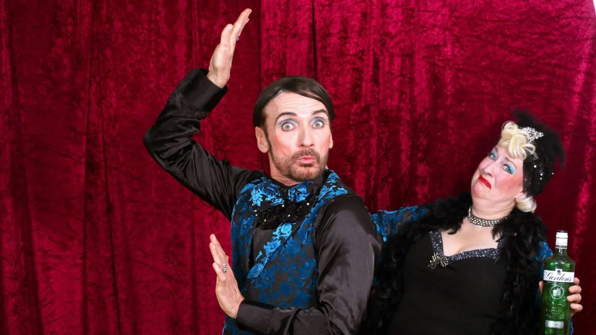A man and a woman in front of a red curtain smile and pose whilst wearing sequinned clothes.