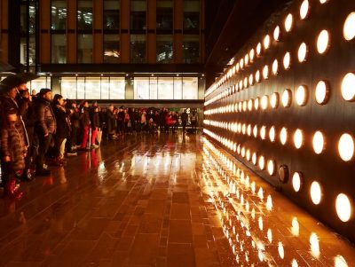 Crowd gathered in the rain faces a wall of glowing orange circular lights, reflecting on the wet pavement. The scene conveys a mood of awe and anticipation.