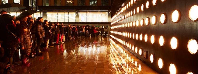 Crowd gathered in the rain faces a wall of glowing orange circular lights, reflecting on the wet pavement. The scene conveys a mood of awe and anticipation.