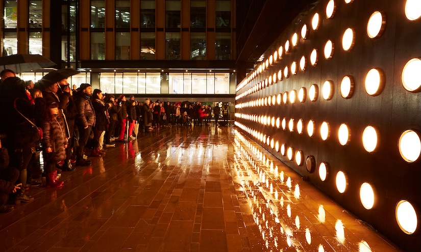 Crowd gathered in the rain faces a wall of glowing orange circular lights, reflecting on the wet pavement. The scene conveys a mood of awe and anticipation.