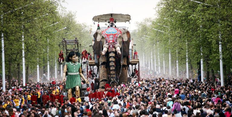 A giant elephant and girl puppet walking down the London Mall. Huge crowds of people are seen around them.