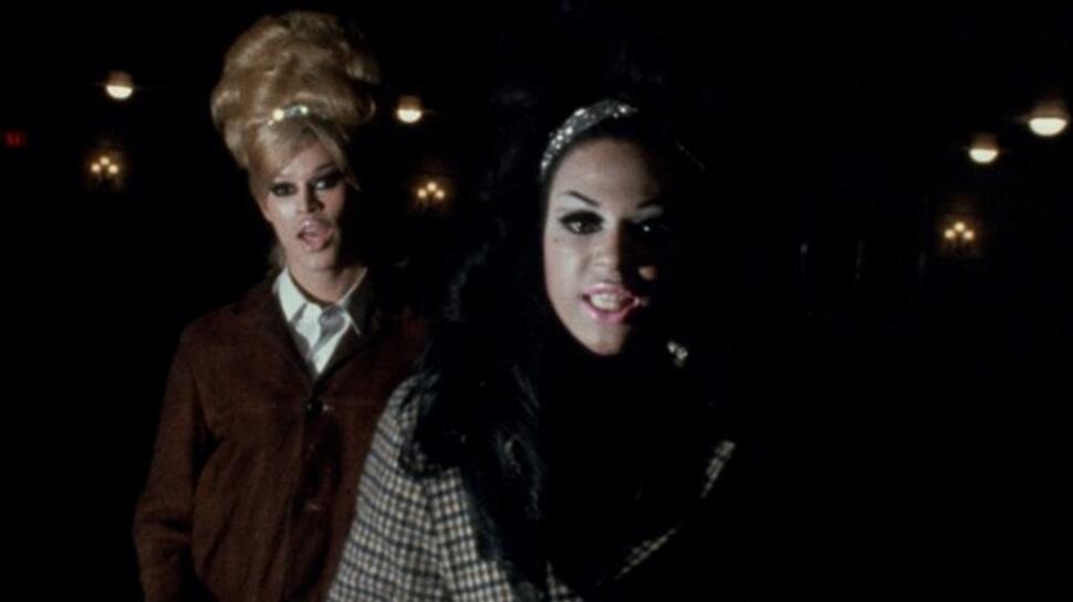 Two drag queens walk a street at night, wearing coats and sparkly headbands.