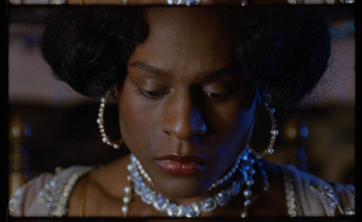 Close up of a Black woman in pearls and regency wear looks downwards of the camera.