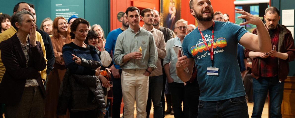 A gallery filled with people whilst a man in a blue shirt presents at the front.