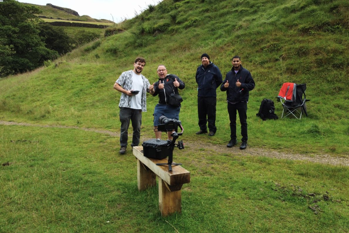 Four members of the video production crew posing and giving thumbs up on a grassy hillside in the Yorkshire countryside, with camera equipment on a wooden bench in the foreground.