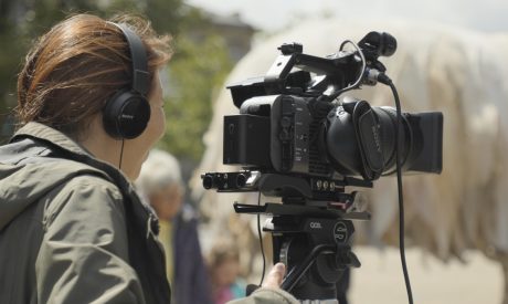 Close-up of a videographer wearing Sony headphones operating a professional Sony camera on a tripod, seen from behind, with a blurred crowd in the background.