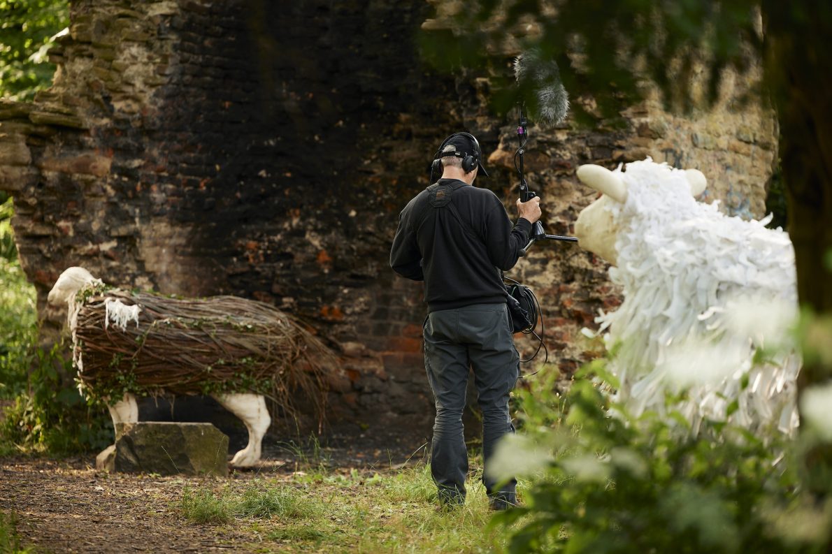 A sound recordist wearing headphones holds a boom microphone between two large sheep puppets — one made from wicker and foliage, one from white fabric — against a crumbling brick wall in a woodland setting at Thornhill Rectory Park.