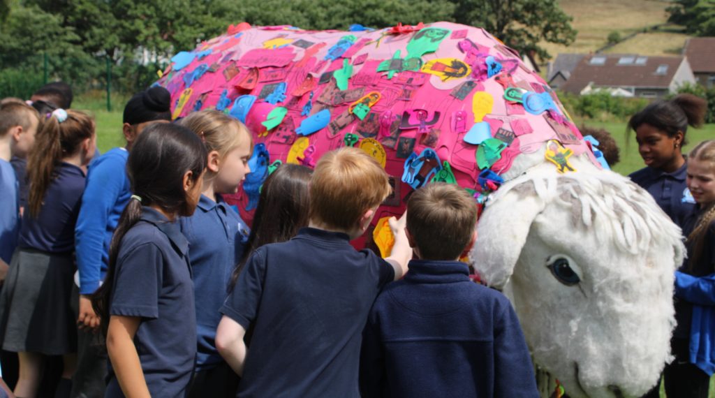 Schoolchildren pet and interact with a large sheep sculpture in a field.