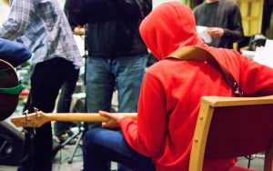 Image of a young person in a red hoodie playing a guitar whilst sat in a music studio