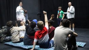 A group of children raise their hands and participate in a workshop in a dark theatre room