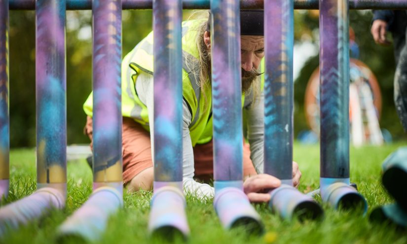 A man diligently adjusting a pipe joint from behind a sculpture. The artwork is a musical instrument made out of recycled pipes and various other junkyard scraps. The pipes are colourfully spray painted in blues and purple.