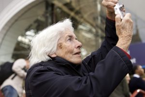 An elderly woman holds up a digital camera to take a picture of something off screen outside of Liverpool Lime Street station.
