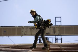 A lone worker in a hard hat and full harness walks with assured footing along the edge of a rooftop structure against a clear blue sky, tools hanging from their belt.