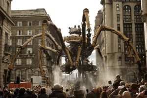 The mechanical spider moves through a packed Liverpool street in daylight, jets of water spraying from its body as it towers above the crowd between grand Edwardian stone facades, operators visible riding its superstructure above the sea of onlookers below.