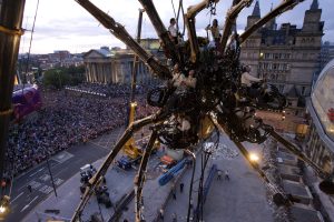 A dramatic aerial view of the mechanical spider suspended high above Liverpool city centre at dusk, its operators clinging to its body and legs, with an enormous crowd stretching back towards St George's Hall and the city skyline beyond, the sheer scale of both the sculpture and the audience laid bare from above.