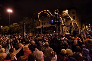 The mechanical spider looms over a vast crowd of spectators on a Liverpool street at night, hundreds of people holding up cameras and mobile phones to capture the moment, the spider's illuminated body and legs rising above the rooftops of the surrounding buildings.