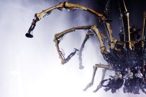 A close-up of the mechanical spider's legs and underbelly shrouded in mist and water spray, the intricate tangle of bone-like limbs, cables, and hydraulic components filling the frame, with operators just visible clinging to the machinery in the lower right corner.