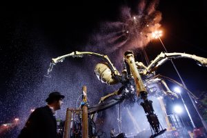 A low-angle night shot of the mechanical spider looming overhead, its yellow bone-like legs spanning the frame and its masked head tilting downward, surrounded by a haze of water spray and a burst of orange smoke, with a silhouetted figure in the foreground and floodlights blazing behind.