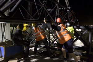 A crew of workers in hi-vis jackets strain together to manoeuvre a large steel structure in the dark, their hands pressed against the framework in a coordinated push.