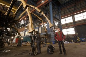 Two people stand dwarfed beneath the mechanical spider's vast bone-like leg structure inside a large industrial warehouse, the scale of the machinery dramatically apparent against the high ceiling and steel roof trusses above.
