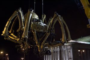 The mechanical spider hangs suspended by crane cables against the night sky, its full form visible with all eight bone-like legs curled beneath its domed body, the floodlit colonnade of St George's Hall glowing behind it and a yellow crane arm visible in the upper right corner.