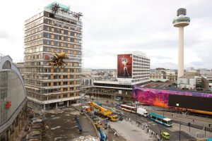 La Machine's giant golden mechanical spider clings to the side of a scaffolded office tower in Liverpool city centre, with buses, cranes and the Radio City tower behind it under an overcast sky.