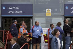 A crowd of onlookers, including Royal Mail workers in light blue uniforms carrying red mail bags, stand outside the Liverpool Lime Street station entrance, all gazing upwards at La Machine.