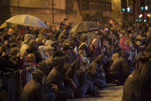 large crowd of spectators packed behind barriers on a wet Liverpool street at night, some sheltering under umbrellas and speckled with what appears to be falling foam or snow, their faces rapt with attention as they watch an event offscreen.
