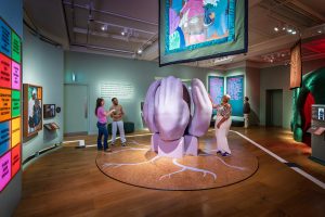Inside a gallery space exploring themes of grief and care, a large sculptural installation of two giant lilac hands, cupped together as if in comfort, rises from a plinth on a circular cork platform painted with white root-like branches radiating across the wood floor around it. A woman in white touches one of the sculpture's hands, while a man and a woman stand talking nearby beside a green bench seat. Above, a large banner hangs from the ceiling, illustrated in a folk-art style with two figures at a table arranging flowers. On the left, a curved wall displays a quote from Camille Sapara Barton reflecting on the value of collective, community-based grieving spaces, above a colourful grid of tiles naming different forms of support. Beside it hangs a folk-art illustration of two embracing figures with accompanying wall text. On the right, backlit teal and pink panels display further exhibition text on community-led care and the long-term impact of growing up in foster care, near a doorway where two more visitors look towards a green and red sculptural form with its own text banner.