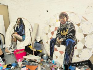 Two large circular mosaic portraits made from fragments of mirror and coloured glass or tile, propped against a white brick studio wall. On the left, a smaller circular artwork shows a seated woman with long dark hair, resting her chin on her hand, wearing a blue top and shorts with white trainers, set within a geometric mosaic border of cream, grey and pale gold triangles. On the right, a larger circular artwork shows a seated man wearing a black jumper with a gold Batman logo, dark trousers and blue-cuffed boots, one arm resting on his knee, surrounded by a similar mosaic border in cream, bronze and grey tones. Between and around the two artworks, the studio floor is cluttered with the artist's working materials: a pink plastic tub and a black crate holding sheets of mirror and glass, a clear plastic storage box, a metal paint tin, scattered offcuts and shards of coloured material, and other supplies in trays and baskets across the concrete floor.