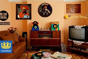 A recreated Caribbean-British front room decorated in warm peach and terracotta tones, with a shag-pile rug underfoot. On the back wall, a square gilt-edged frame holds a photograph of a woman in an orange headwrap and jumpsuit, headphones round her neck, holding a record sleeve that reads "Otis Gayle". Beside it, a circular frame holds a photograph of a man in a red, gold and green knitted hat, singing into a microphone against a starry backdrop. To the right, a carved wooden wall plaque shows a map of the Caribbean islands labelled "CARIBBEAN", and below it an illustrated figure in a headwrap dances beside the words "Nzinga Dance" in yellow script. In the centre of the room stands a dark wood radiogram, branded "Blue Spot", topped with a stand of colourful vinyl singles and a green record sleeve; further records lean against its side and are stacked on the floor beside it. To the right, an old cathode-ray television sits on a wooden stand draped with a crocheted doily, with a stack of magazines on the shelf below. On the left, a brown sofa with patterned cushions sits beside a small table holding a vase of flowers and framed photographs, with more vinyl records propped against the wall nearby. In the foreground, a low coffee table holds a glass bowl of pink and white sweets, a folded copy of the Jamaican newspaper "Daily Gleaner", and further records. In the top left corner, partly cropped by the edge of the image, a black circular logo reads "B:M 2026" above the partly visible words "honouring our communities". In the bottom left corner, a blue square logo shows a yellow crown above the word "Lewisham".