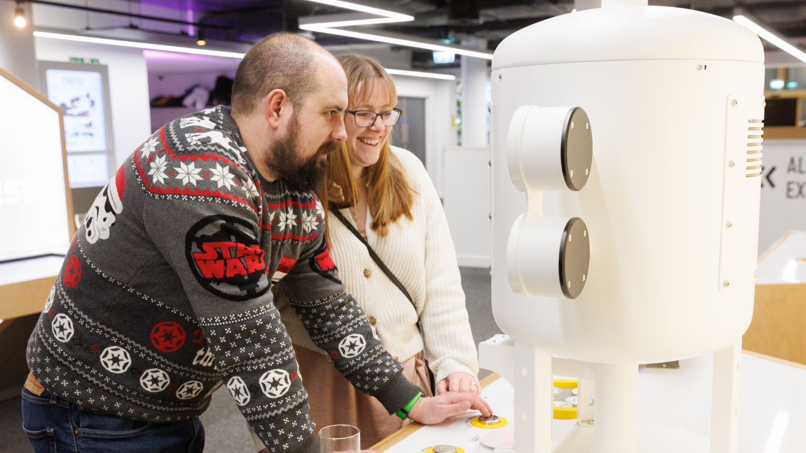 A young couple wearing Christmas jumpers and interacting with a hands-on science exhibit