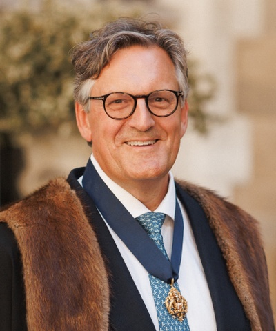 Robert looks straight-on into the camera wearing a pale blue shirt and the MT badge with a faux-fur trimmed black robe