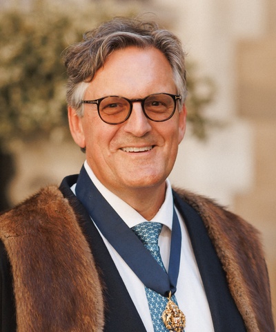 Robert looks straight-on into the camera wearing a pale blue shirt and the MT badge with a faux-fur trimmed black robe