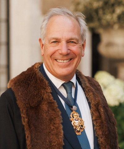 The Master smiles facing the camera wearing a black robe with fake fur brown trim and wearing a badge of the Merchant Taylors' crest