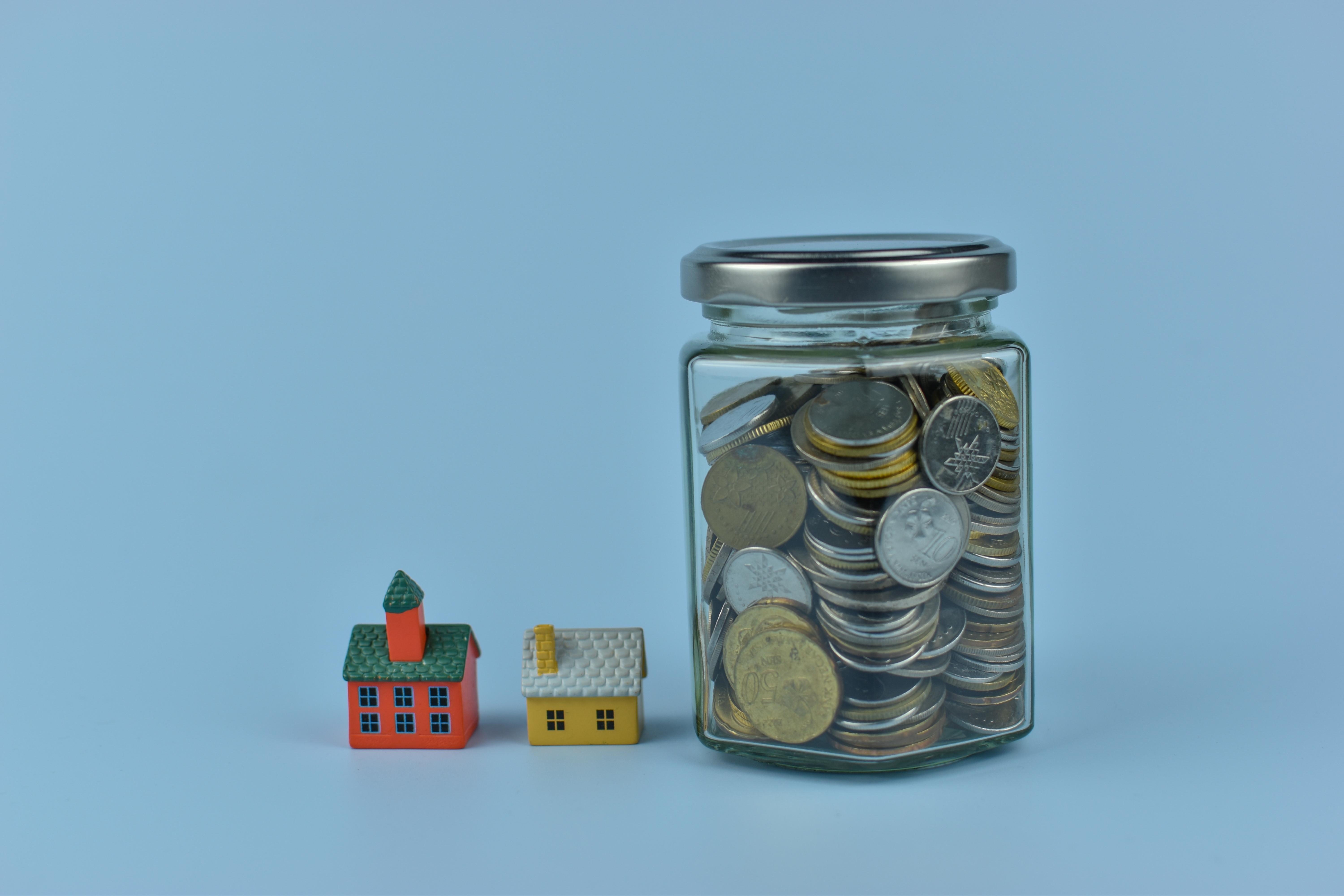 Tiny houses sit beside spilling coins. Glass jar tilts, coins cascade softly. Bright colors contrast with calm blue background. No people, no animals, no plants shown